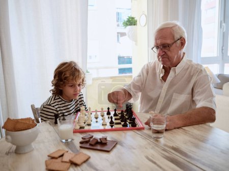Foto de Hombre mayor enseñando niño preadolescente concentrado a jugar ajedrez mientras está sentado en la mesa en la cómoda sala de estar en casa - Imagen libre de derechos