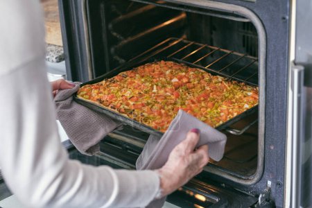 Foto de Mujer anciana irreconocible en proceso de cocción poniendo pizza vegetal en bandeja para hornear en el horno abierto en la cocina - Imagen libre de derechos