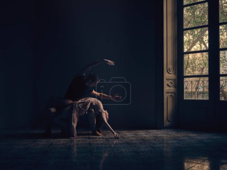 Foto de Cuerpo completo de bailarina flexible acostada sobre las rodillas de un bailarín masculino mientras realiza la danza en un amplio estudio con poca luz - Imagen libre de derechos