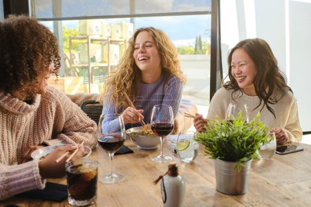 Foto de Mujeres jóvenes y diversas satisfechas mirándose mientras comen ramen en la mesa de madera cerca de copas de vino tinto - Imagen libre de derechos