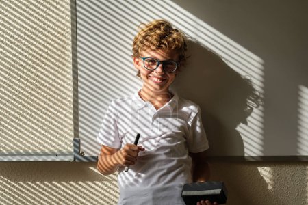 Foto de Deleitado niño en gafas de pie con marcador y borrador cerca de pizarra en el aula y mirando a la cámara - Imagen libre de derechos