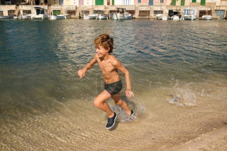 Téléchargez les photos : Full body side view of shirtless tanné preteen child rire heureux tout en courant dans l'eau de la baie de la mer dans la ville balnéaire côtière en été - en image libre de droit