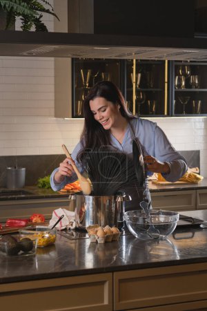 Photo for Satisfied lady holding lid and mixing black pasta in saucepan with wooden spatula while standing near kitchen counter - Royalty Free Image