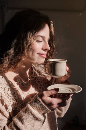Téléchargez les photos : Vue latérale d'une jeune femme blonde aux cheveux bouclés dégustant une tasse de café chaud dans une pièce lumineuse - en image libre de droit