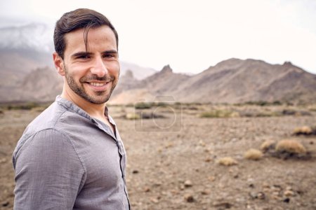 Photo for Cheerful bearded male traveler in casual clothes standing on dry grassy stony arid terrain and smiling looking at camera - Royalty Free Image
