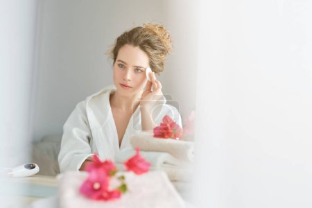Téléchargez les photos : Charmante femme en peignoir essuyant le visage avec du coton tout en regardant miroir près de la table avec des serviettes pendant la routine du matin - en image libre de droit