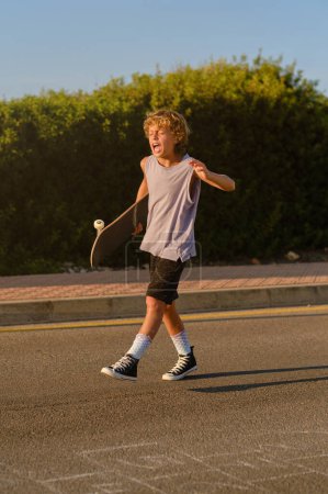 Photo for Excited preteen hipster with skateboard walking along road and screaming while enjoying summer evening - Royalty Free Image