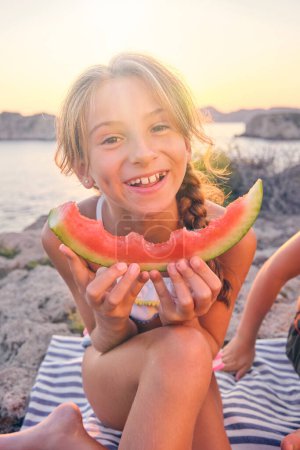 Photo for Smiling preteen child in swimsuit having fun while eating delicious watermelon and sitting on striped picnic blanket on summer evening - Royalty Free Image