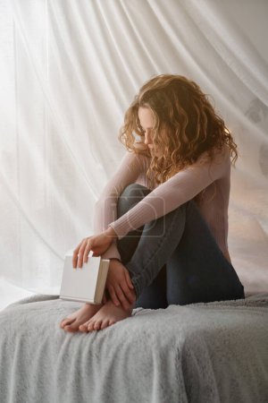 Foto de Encantadora joven con ropa casual sentada en una cama acogedora con libro cerca de la cortina blanca y abrazando las rodillas en el dormitorio moderno - Imagen libre de derechos