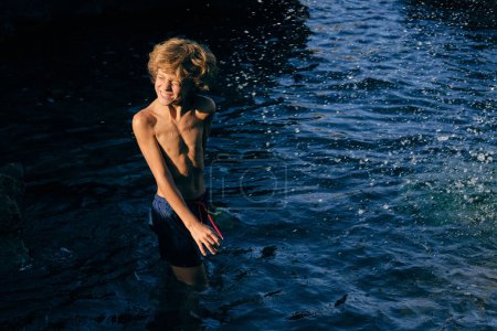 Téléchargez les photos : Enfant dans des troncs de natation les yeux fermés faisant face à l'océan avec éclaboussures d'eau au soleil - en image libre de droit