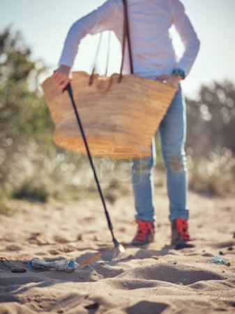 Téléchargez les photos : Cultivez un garçon préadolescent anonyme avec un sac en osier à la recherche de déchets plastiques et de déchets avec ramasse-ordures sur une plage de sable fin par temps ensoleillé - en image libre de droit