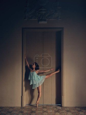 Foto de Vista lateral de la señora flexible en vestido de verano y zapatos puntiagudos practicando arabesco contra la puerta de la habitación en luz tenue - Imagen libre de derechos