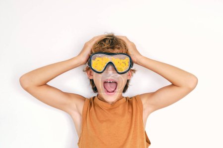 Téléchargez les photos : Enfant insouciant avec bouche ouverte dans un masque de plongée avec des algues jaunes criant sur fond blanc - en image libre de droit