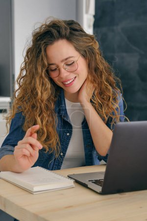 Photo for Young female entrepreneur smiling and looking at notepad while working on project remotely using netbook and touching neck at home - Royalty Free Image
