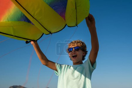 Photo for Low angle of delighted curly haired preteen child in sunglasses enjoying summer holidays while holding multicolored kite against blue sky - Royalty Free Image