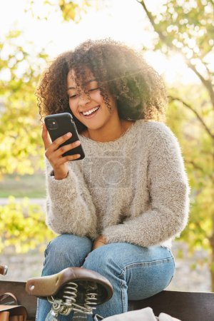 Foto de Mujer hispana sonriente con el pelo rizado en suéter cálido y vaqueros sentados en un banco de madera cerca de la bicicleta y mensaje de texto a través de teléfono inteligente durante el fin de semana en el parque en el día soleado de otoño - Imagen libre de derechos