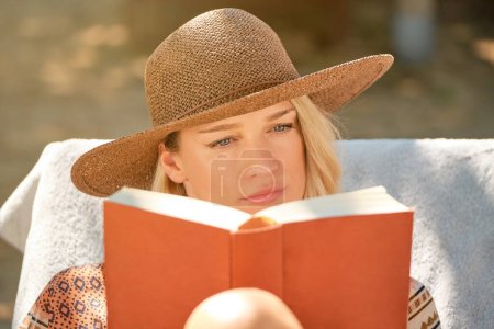 Téléchargez les photos : Jeune femme pensive en chapeau et veste de paille regardant le livre borné de rouge tout en lisant avec concentration et assise sur un matelas blanc sur un transat - en image libre de droit