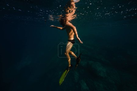 Foto de Vista lateral cuerpo completo de buzo masculino irreconocible sin camisa en aletas y gafas flotando bajo el agua azul del mar profundo en el día soleado - Imagen libre de derechos