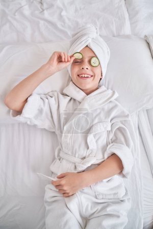 Foto de Alto ángulo de sonriente niño con turbante toalla demostrando rodaja de pepino mientras descansa en la cama blanca durante los procedimientos de spa en el dormitorio en casa - Imagen libre de derechos
