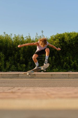 Foto de Nivel del suelo de niño preadolescente fresco saltando con monopatín sobre la carretera y mostrando truco en la ciudad por la noche - Imagen libre de derechos