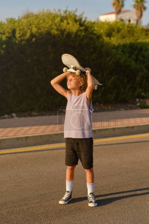 Photo for Serene preteen skater standing on road with skateboard above head and enjoying summer sunset with closed eyes - Royalty Free Image