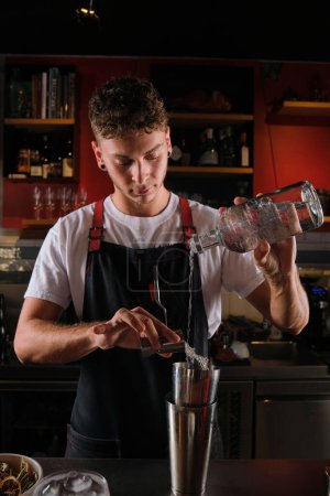Téléchargez les photos : Jeune barman mâle avec des boucles d'oreilles debout près du comptoir et verser de la vodka de la bouteille et du sirop de jigger dans un shaker de cordonnier en métal tout en préparant un cocktail - en image libre de droit