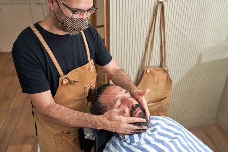 Téléchargez les photos : Coiffure masculine professionnelle dans le masque de protection massage menton du client pendant la procédure de traitement de la barbe dans le salon de coiffure moderne - en image libre de droit