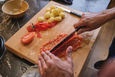 Foto de Hombre de la cosecha picando tomate rojo cuidadosamente en el tablero de madera con la mentira peladas pequeñas papas en la cocina - Imagen libre de derechos
