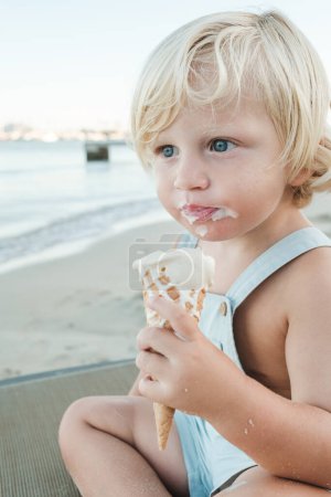 Foto de Niño adorable con el pelo rubio y la boca desordenada mirando hacia otro lado mientras está sentado en la orilla del mar y disfrutando de delicioso cono de helado - Imagen libre de derechos