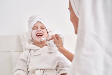 Photo for Mother applying moisturizing facial mask with brush on laughing kid in bathrobe and towel on head sitting on bed and leaning on pillows while enjoying spa procedure - Royalty Free Image