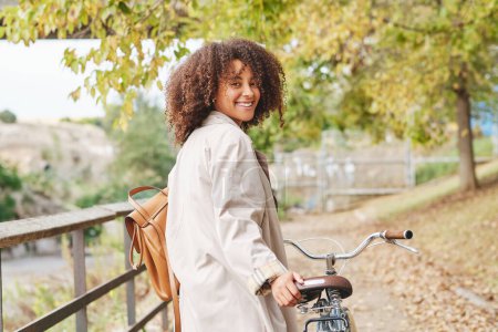 Téléchargez les photos : Vue latérale de la jeune femme afro-américaine avec des cheveux bouclés dans un manteau élégant debout avec vélo et regardant la caméra dans le parc - en image libre de droit