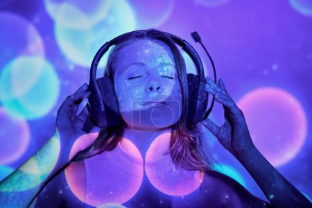 Foto de Mujer joven ajustando auriculares y disfrutando de buena música bajo la proyección de destellos de colores - Imagen libre de derechos