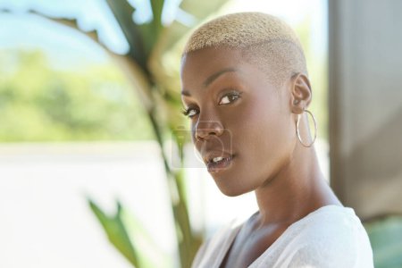 Foto de Retrato de vista lateral de la joven afroamericana con estilo reflexivo con el pelo corto y rubio y en pendientes de oro mirando a la cámara - Imagen libre de derechos