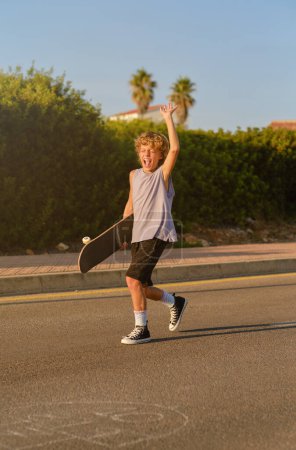 Photo for Cheerful preteen skater with skateboard walking with raised arm on road and enjoying summer weekend while screaming with closed eyes - Royalty Free Image