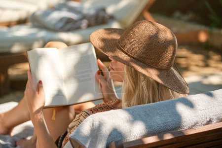 Photo for Side view of young female barefoot traveler with hat sitting on wooden bench and reading book while looking away at resort in sunlight outdoors - Royalty Free Image