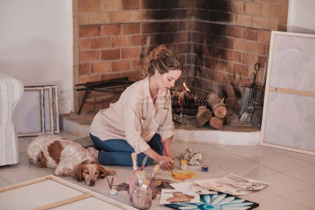Photo for Side view of concentrated female artist in casual clothes sitting near fireplace and drawing on fabric using paintbrush and professional paint and pet sleeping near in creative workspace - Royalty Free Image