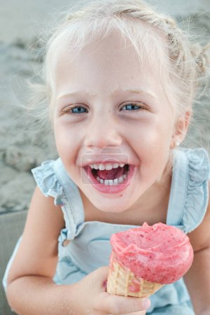 Photo for High angle of blond kid looking at camera and laughing while eating delicious ice cream on blurred background of beach - Royalty Free Image