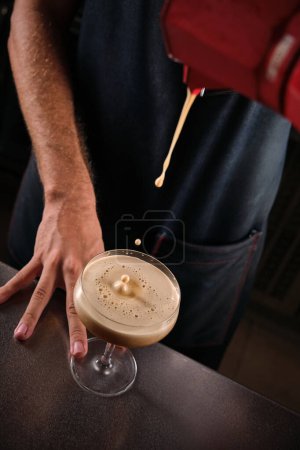 Photo for From above of crop unrecognizable male barkeeper in black apron standing near counter and pouring fresh coffee cocktail from moka pot into champagne coupe in bar - Royalty Free Image