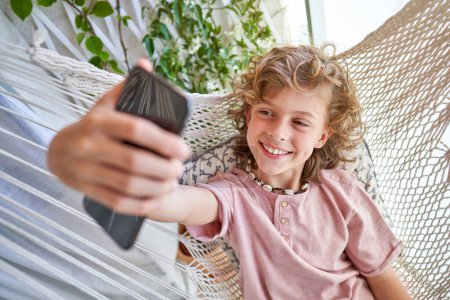 Photo for Smiling preteen boy with curly hair in pink t shirt taking selfie on smartphone while resting in wicker hammock on balcony - Royalty Free Image