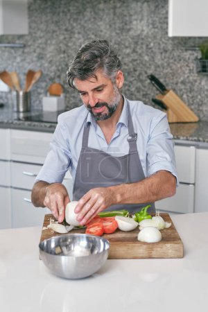 Photo for Bearded male cook in apron peeling onion on cutting board with fresh vegetable while cooking at table in light kitchen - Royalty Free Image