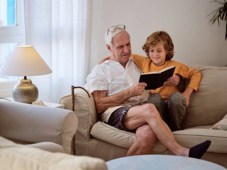 Photo for Senior man reading book with content grandson while chilling together on comfortable sofa in cozy living room - Royalty Free Image
