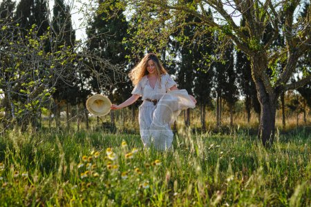 Foto de Longitud completa de la mujer sensible con maxi vestido blanco paseando por el verde prado herboso en el exuberante parque de verano - Imagen libre de derechos