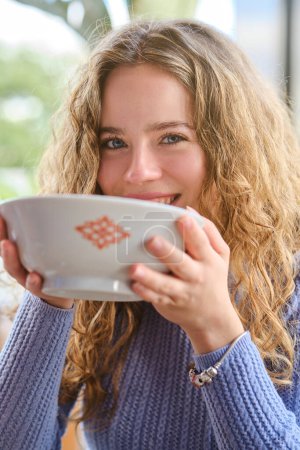 Photo for Charming curly haired woman in knitted sweater looking at camera and drinking ramen soup from ceramic bowl while spending time in Asian restaurant - Royalty Free Image