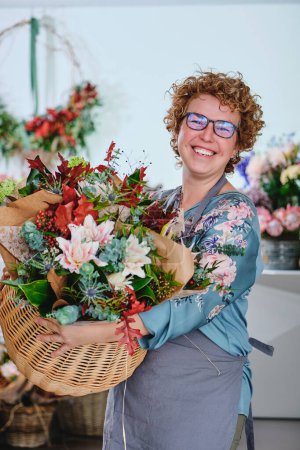 Foto de Florista femenina positiva en delantal y gafas con ramo de flores en cesta sonriendo y mirando a cámara en tienda floral - Imagen libre de derechos