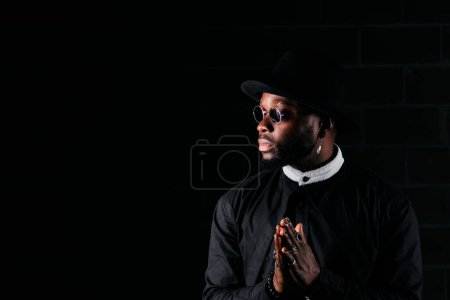 Foto de Modelo masculino afroamericano serio con manos de oración que usan ropa negra de moda mirando hacia otro lado sobre fondo oscuro - Imagen libre de derechos