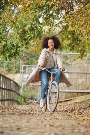 Foto de Cuerpo completo de mujer afroamericana feliz en ropa casual montar en bicicleta mientras se divierten en el parque de otoño - Imagen libre de derechos