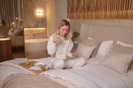 Téléchargez les photos : Femme souriante en pyjama blanc assis sur le lit avec les jambes croisées et surfant téléphone mobile tout en prenant le petit déjeuner dans la chambre à coucher à la maison le week-end - en image libre de droit