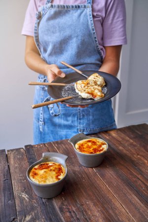 Photo for Unrecognizable female in denim apron standing with plate of roasted pastry in chopsticks at wooden counter while preparing bowls of fried food for serving - Royalty Free Image