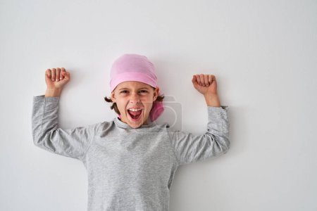 Foto de Niño alegre en camisa gris y pañuelo rosa apretando puños y gritando fuerte mientras se apoya en la pared blanca y mirando a la cámara durante la recuperación en la clínica - Imagen libre de derechos