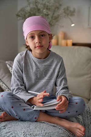 Téléchargez les photos : Garçon en tenue de nuit et foulard rose pour sensibiliser le public au cancer du sein assis avec les jambes croisées et écrire dans un bloc-notes tout en regardant la caméra - en image libre de droit